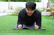 © 220 Selfmade studio - Fit Asian man doing plank exercise black yoga mat outdoors backyard garden feeling determined focused and strong during morning workout routine