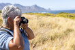 © Wavebreak Media - Senior woman holding DSLR camera capturing coastal grasses with mountains over ocean, copy space
