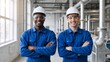 © Vasiliy - Two smiling industrial workers in blue jumpsuits and hard hats. Diverse team of engineers standing with arms crossed in a factory. Professional workplace safety and teamwork concept