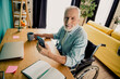 © deagreez - Senior man in wheelchair at home office smiling while using smartphone beside laptop and notebook in bright living room scene
