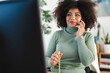 © Studio Marmellata - A woman with curly hair talks on her phone while holding a pencil, working at her desk with a computer in front of her.