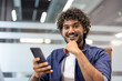 © Tetiana - Close-up portrait of a young Indian man sitting in a chair at a desk in an office, holding a phone and smiling at the camera