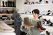 © JackF - Female customer is standing next to shelf in stores sales area. She picks up shoes, looks at and studies material and quality of product, decides to buy shoes