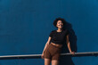 © BUDDHA - Smiling young woman with brown shorts and black t-shirt leaning on a railing in front of a blue wall