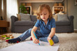 © Stockphotodirectors - A young child sits on a soft carpet, focused on drawing with a blue pencil and a yellow sponge.