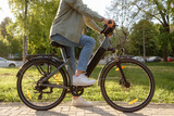 Bicyclist rides electric bike in park during sunny day with green trees around in early afternoon