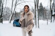 © standret - Woman in winter attire, smiling warmly while standing in a snowy landscape, surrounded by trees, showcasing a cozy and stylish winter fashion look with a vehicle in the background