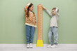 © New Africa - Little boy and girl measuring their height with chart near light green wall
