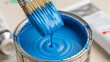 © Aline Shawn - A close-up, high-angle shot focuses on a metal paint can filled with vibrant, bright blue liquid paint. A paintbrush, held from above, is dipped into the paint.