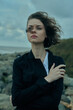 © SHOTPRIME STUDIO - Moody woman on the coast, wind in hair, outdoors scene with sea, rocks and cloudy sky, wearing a dark jacket, intimate yet distant pose, cinematic mood for fashion, lifestyle, nature, travel