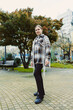 © DSMT - Young woman stands in park wearing a checkered coat with fringe, surrounded by trees and benches during a cloudy day