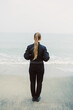 © DSMT - Person stands on sandy beach near calm water with back turned while waves gently touch the shore during a cloudy day in coastal area
