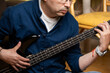 © Claudio Bórquez - Cropped shot of man playing black electric bass guitar while sitting on floor, Close up of musician hands on four string bass guitar neck during home rehearsal