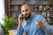 © Liubomir - Young bald man making phone call, experiencing communication issues and expressing frustration or misunderstanding while teleworking from a home office environment
