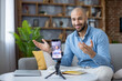 © Liubomir - Young bald bearded man smiling and speaking to camera while recording a live stream or online webinar with smartphone on tripod in a casual home living room setting