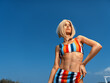 © SHOTPRIME STUDIO - confident young woman wearing colorful striped bikini standing outdoors against clear blue sky, looking away with hand on hip, summer beachwear style
