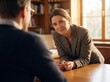 © Olha - Professional middle-aged woman leaning forward on a desk, listening empathetically to a client during a consultation in a sunlit office with bookshelves.
