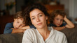© forenna - Tired mother resting on a sofa while children play nearby, heavy eyes and stillness capturing fatigue, devotion, and everyday family reality. cinematic color correction, natural uneven lighting yet