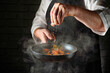 © Віталій Б. - A chef in an apron prepares a dish, sautйing chopped vegetables in a frying pan. The cook adds salt as steam rises from the pan in a busy kitchen