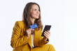 © Alliance - A smiling professional woman in a yellow blazer confidently holds a credit card and smartphone, symbolizing seamless online payments and modern financial convenience.
