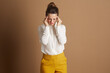 © Alliance - A professional woman in a white shirt and yellow trousers grimaces in pain, holding her temples, suggesting a stress headache or discomfort in a corporate setting.