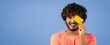 © Prostock-studio - A young man with curly hair holds a yellow card while smiling. He stands against a blue background and wears a light-colored shirt. The scene takes place during daytime hours.
