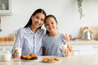 © Prostock-studio - Young asian mother and daughter enjoying fresh homemade cookies, drinking milk and smiling to camera while sitting at table in kitchen, spending time together at home