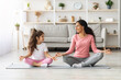 © Prostock-studio - Cheerful young mother and little daughter in sportswear sitting on fitness mat in lotus position and smiling to each other, meditating together at home, copy space. Healthy family, sports lifestyle