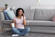 © Prostock-studio - Free time, rest and calls to friend on phone, coffee break at home. Satisfied millennial pretty african american woman talking on smartphone, hold cup of hot drink in minimal living room interior