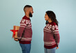 © Prostock-studio - Surprise for Christmas. Black guy holding gift box behind back, congratulating wife over blue studio background. Happy african american couple in sweaters celebrating xmas