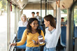 © Prostock-studio - Portrait of two happy young multiethnic ladies standing inside bus and chatting, drinking coffee cup and using cellphone technology, friends commuting together to work or high school university