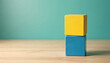 © Pete - Yellow block stacks on blue block. Two colorful wooden cubes balance on light wood table. Minimalist concept on teal background. Abstract arrangement offers metaphor for choice, balance, idea,