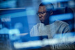 © Seventyfour - Black man young adult sitting at desk working on computer monitor in modern office, wearing glasses and focusing on screen, technology reflections visible in foreground