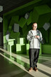 © Seventyfour - Young adult Caucasian man standing indoors using smartphone, looking down at device with neutral expression, geometric green abstract wall and cubes in background