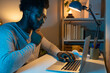 © Daniel - Pensive African American male college student studying late at night at home. Black man working using laptop.