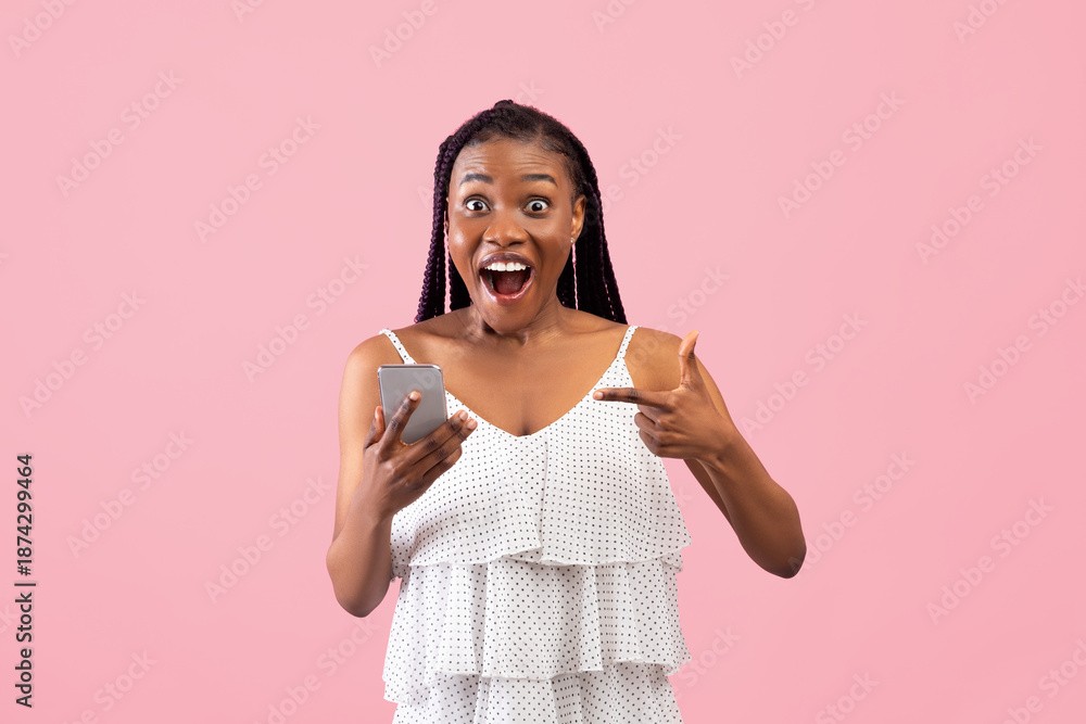 Shocked black lady pointing at smartphone in excitement, winning lottery or online casino bet on pink studio background. Excited African American woman receiving amazing news on phone
