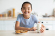 © Prostock-studio - Portrait of happy asian girl enjoying cookies and milk, sitting in kitchen and smiling to camera. Pretty child eating fresh baked biscuits and drinking calcium drink, free space