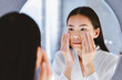 © Prostock-studio - A woman stands in front of a mirror in a bathroom. She gently applies skincare products to her face with both hands. The setting is bright and features a modern design.