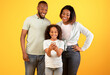 © Prostock-studio - Parenting control apps concept. Little black girl holding smartphone and parents looking at cellphone, standing together over yellow background, studio shot