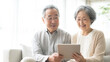 © SHOKO - ソファで並んでタブレットを見て楽しむ笑顔のシニア夫婦