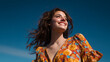 © Nova Zigres - Joyful young woman photographed from a low angle, brown hair moving slightly in the wind, bright blue sky, optimistic lifestyle mood.