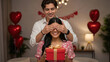 © ArtFocus Studio - Smiling young man surprises his girlfriend with a red gift box while covering her eyes in a room decorated with heart balloons for Valentine's Day.
