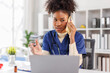 © David - Happy friendly young woman doctor, nurse, medical worker, reception specialist in blue uniform with stethoscope sitting at desk, talking to patient on phone and checking schedule information on laptop