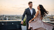 © Vyatcheslav - Couple dancing on a rooftop at sunset. Man in formal suit and woman in flowing gown celebrating in the city. Romantic date night with urban skyline view. Luxury lifestyle and relationship concept