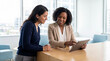 © Таня Андреева - Two diverse businesswomen collaborating and smiling while looking at a tablet in a modern, bright corporate office with a large window.