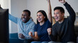 © Таня Андреева - A diverse group of three excited young friends sitting on a couch at home, cheering and celebrating while watching a sports game on TV.