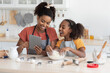 © Prostock-studio - Happy black mummy and daughter cooking at home together, wearing aprons, standing by kitchen table with dough, flour, rolling pin and cookies figurines, using tablet, reading culinary blog