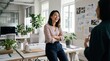 © ArtMajestic - Asian woman leaning against desk and laughing with colleague in bright creative office. Business teamwork and happy corporate culture banner with copy space