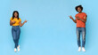 © Prostock-studio - Beautiful millennnial black man and woman in casual outfits showing copy space between them on blue studio background, pointing at advertisement and smiling, full length shot