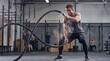 © crispy - Man exercising with battle rope in gym, creating dust. Fitness training and determination concept for health and wellness. High intensity workout